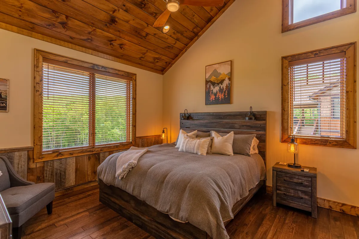 A king bedroom with a plush bed dressed in crisp linens, large windows with blinds allowing in natural light, and calming wooden wall decor. The serene setup invites relaxation and quiet moments.