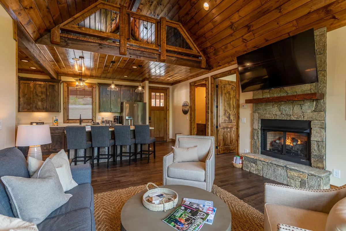 This image showcases a warm, inviting open-plan area featuring a prominent fireplace, a kitchen island, plush seating, and exposed wooden beams. The seamless integration of the living and kitchen spaces is evident, with cozy seating positioned for fireside chats and group gatherings.