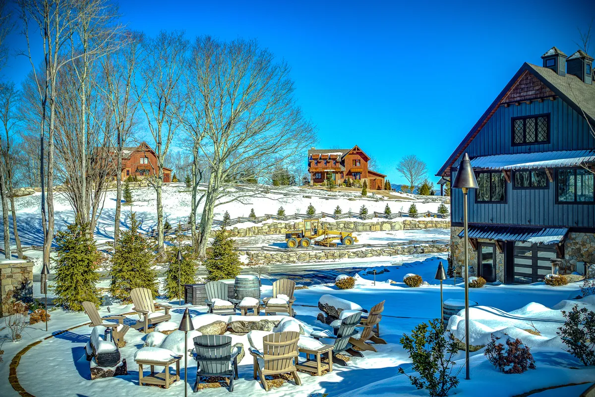 A modern house stands amidst a snowy landscape, with a cozy, snow-covered seating area and fire pit inviting guests to gather outdoors. Nearby, distant cabins dot the wintery scene, framing an atmosphere perfect for relaxing and socializing even in the colder months.
