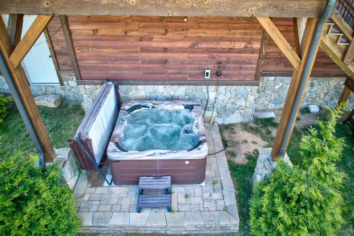 This exterior image features a private hot tub set into a wooden structure and a stone wall, evoking an inviting and luxurious outdoor leisure space. The scene suggests privacy and relaxation, with the hot tub positioned for scenic views and stargazing, ideal for a romantic couple's escape.