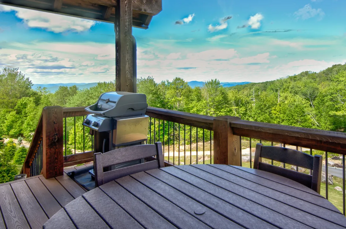 A spacious deck features a grill and a dining table looking out over sweeping mountain views. The inviting setup suggests group barbecues, sunset meals, and outdoor relaxation amid nature's beauty.