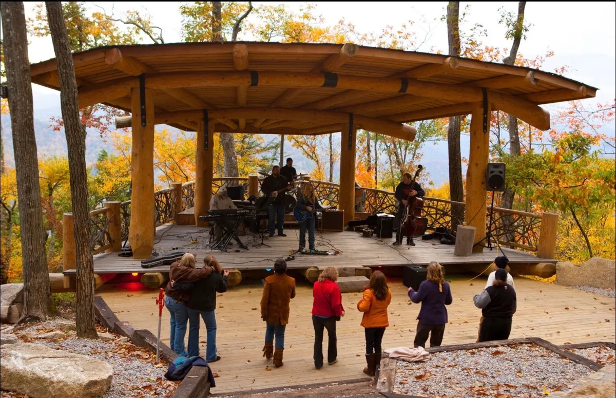An amphitheater scene alive with musicians performing on stage, an enthusiastic audience, and a backdrop of trees colored with fall foliage. The setting conveys a sense of community, leisure, and seasonal festivities.