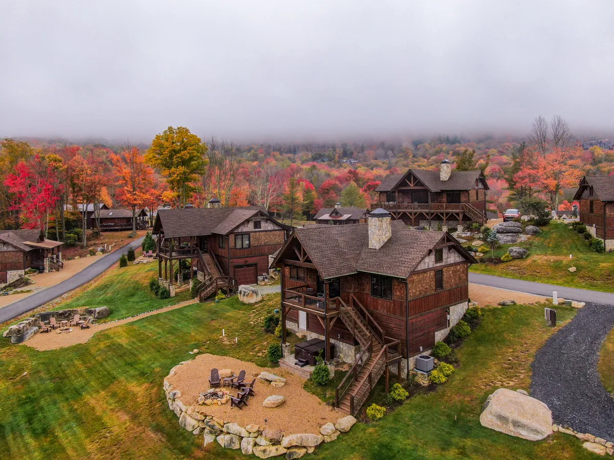 A rustic cabin exterior surrounded by vibrant autumn foliage, featuring a designated fire pit area with seating. The landscape offers a serene backdrop for gatherings, with gravel pathways inviting exploration.
