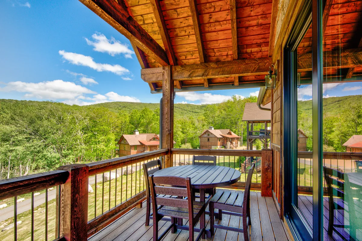 An expansive wooden deck with a stylish outdoor dining set overlooks sweeping mountain vistas. The setting is relaxed yet elevated, perfect for morning coffee, intimate meals, or stargazing. The view stretches over rolling hills and distant peaks, encapsulating the allure of the mountain setting.