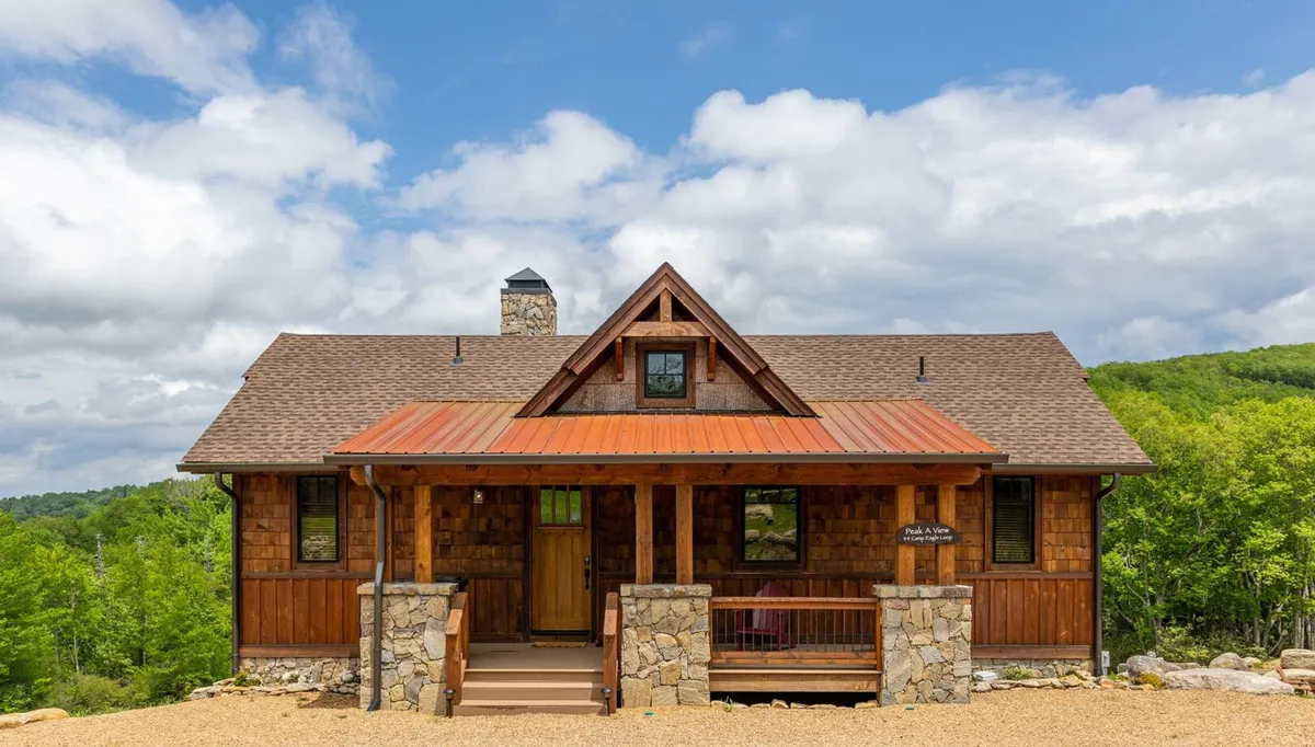 The image showcases a charming wooden cabin with stone accents, nestled in the mountains of Banner Elk. The inviting front porch hints at relaxing mornings or evenings spent with loved ones, while the chimney promises warmth and comfort inside. Dramatic mountain vistas and the soft glow of sunset set a tranquil, welcoming scene.