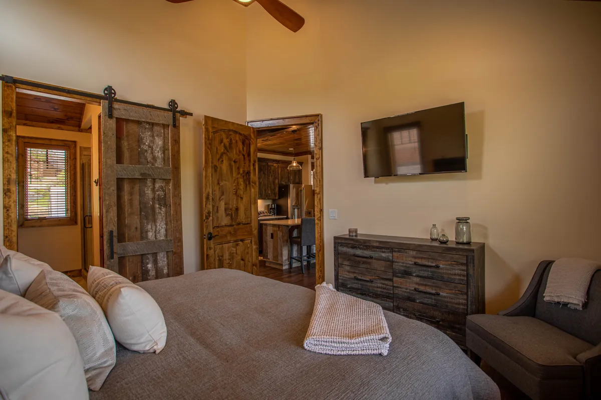 A cozy king bedroom featuring a comfortable bed with decorative pillows, rustic sliding doors, a mounted smart TV, and warm wooden furniture. The space exudes a homely feel, perfect for relaxing and enjoying in-room entertainment.