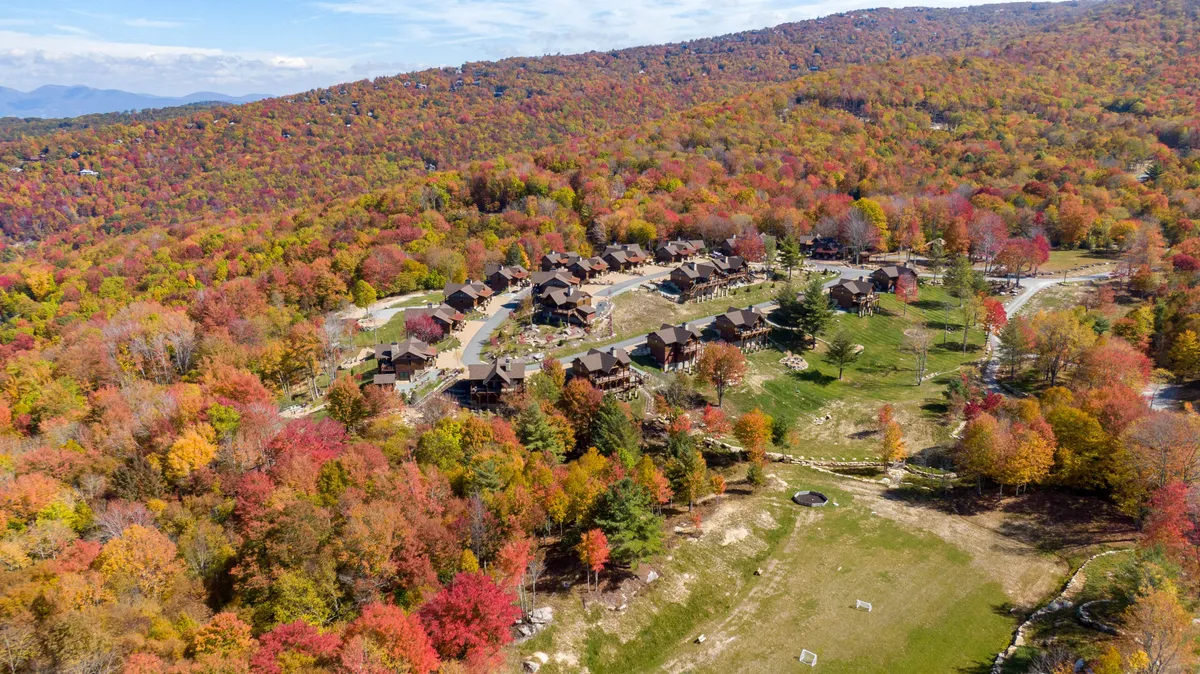 The scene showcases a picturesque residential cluster of cabins surrounded by colorful autumn foliage and a wide, open green space. It's perfect for family gatherings, outdoor games, and exploring nature together.